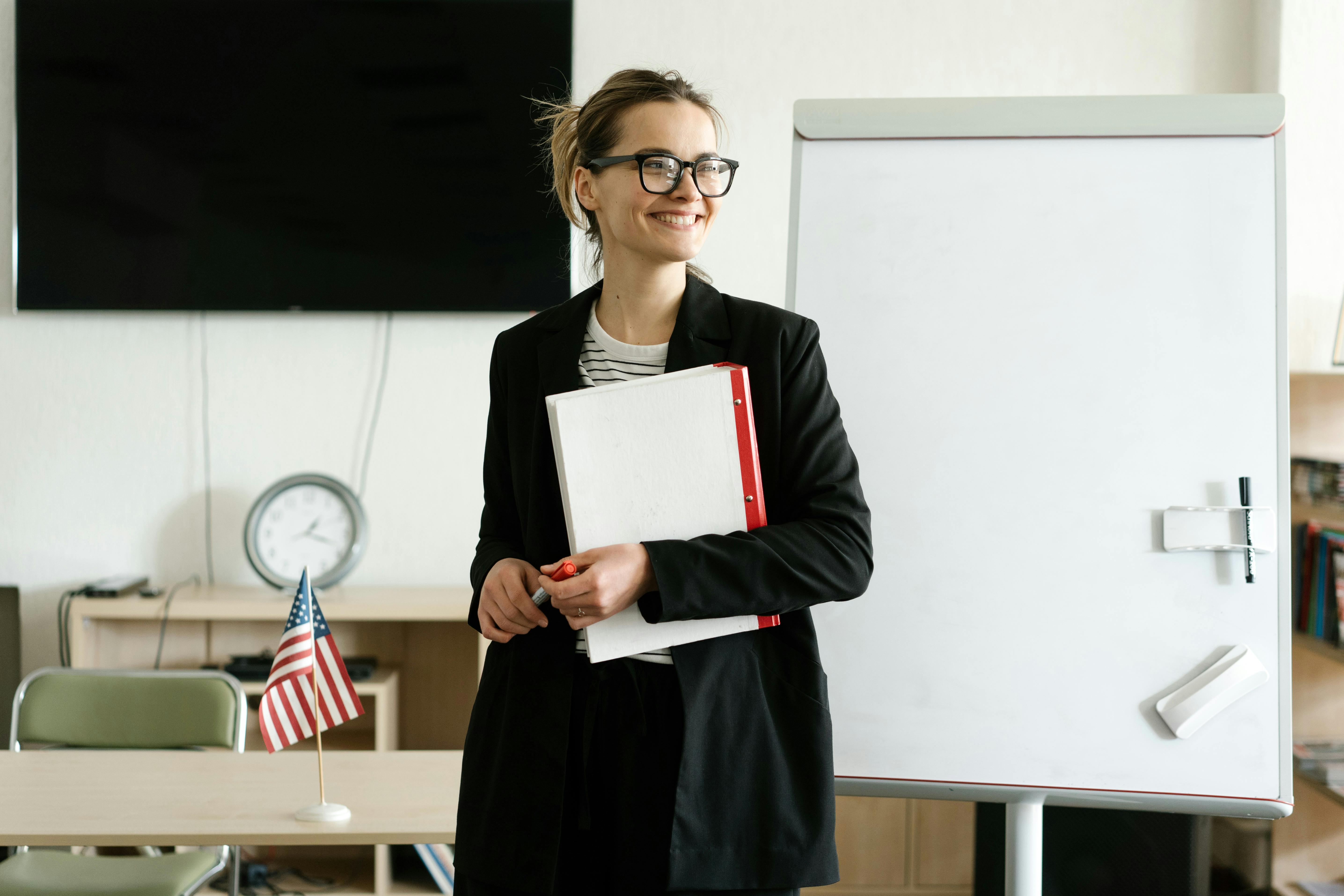 Smiling woman in black blazer holding notebooks next to a whiteboard with a US flag in an office setting.